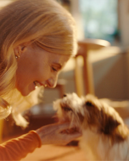 A woman with blonde hair smiles and gently pets a small fluffy dog in a warmly lit indoor setting.